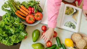 Top view of fresh vegetables and hands arranging them with a recipe book, promoting healthy eating.